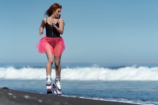 KAMCHATKA, RUSSIA - JUNE 15, 2022: Fitness female in sports Kangoo Jumps boots, black one piece swimsuit and short pink skirt running and jumping on sandy beach during training aerobic session