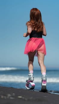KAMCHATKA, RUSSIA - JUNE 15, 2022: Back view of female trainer in sports Kangoo Jumps boots, swimsuit and short pink skirt running and jumping on black sandy beach during fitness, aerobic session