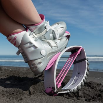 KAMCHATKA, RUSSIA - JUNE 15, 2022: Closeup female legs in Kangoo Jumps shoes on black sandy beach while resting during fitness exercising. Low angle cropped shot of trendy pink sports jumping boots