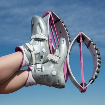 KAMCHATKA, RUSSIA - JUNE 15, 2022: Crop close-up view woman crossed legs up in air shod in pink Kangoo Jumps boots during outdoor aerobic fitness workout on background of blue sky on sunny summer day