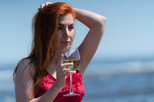 Portrait of redhead woman in red dress holding glass of white wine. Charming woman relaxation on background of blue sky and ocean waves at beach party during summer holidays