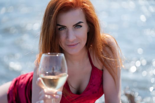 Portrait of happy redhead woman holding glass of wine and looking intently at camera against bokeh background of sea waves at beach party during summer holidays. Sensuality woman in red evening dress