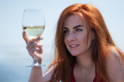 Close-up portrait happy redhead woman has raised glass of white wine at eye level and is looking intently at it. Smiling female in red dress drinking wine on beach on sunny day during summer holidays