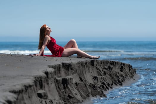 Woman lies leaning on her hands on black sandy beach on steep shore of ocean. Woman in red velvet long party dress with raised hem and bare legs, throws back her head with closed eyes, sunbathes