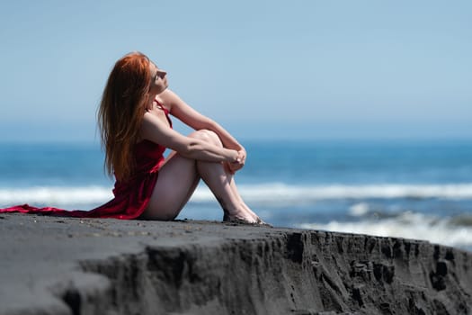 Woman in red velvet long party dress with raised hem and bare legs sitting on black sandy beach, relaxation hugging his knees. Sensuality redhead woman sunbathes throws back her head with closed eyes