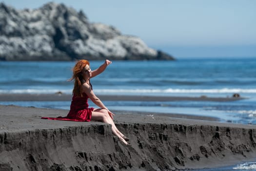 Woman in red long dress with raised hem sitting on sandy beach with bare legs dangling over cliff and shielding herself from summer sun with hand. Elegant redhead fashion model during beach holidays