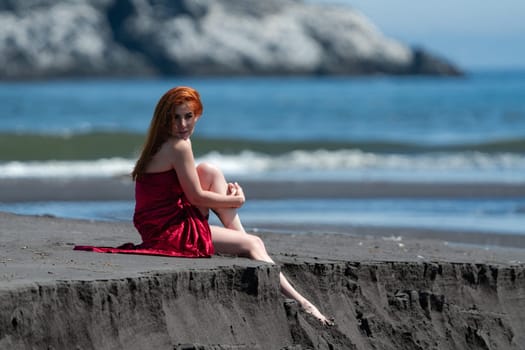 Redhead woman in red long dress with raised hem sitting on sandy beach with bare legs dangling one leg over cliff, tucking other and hugging his arms. Model relaxation during summer beach holidays