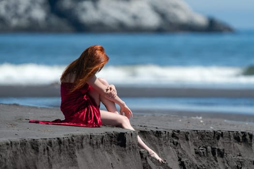 Unrecognizable woman in red dress with raised hem sitting on sandy beach dangling one leg over cliff, and tucking other and hugging his arms. Barefoot female relaxation during summer beach vacations