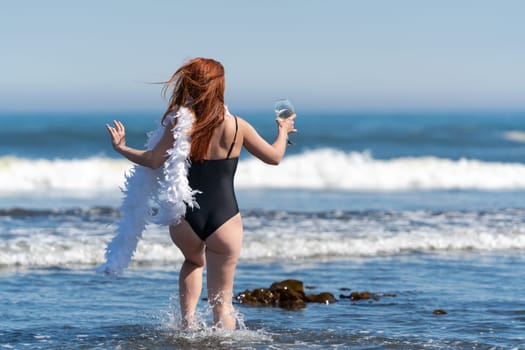 Playful unrecognizable woman in black one piece swimsuit walking ankle deep in water into breaking wave of sea. Woman holding boa in one hand and glass of wine in other. Rear view, full length female