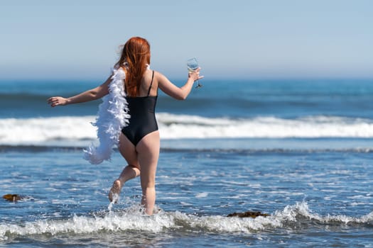 Playful unrecognizable woman walking ankle deep in water into breaking wave of sea. Rear view of woman in black one piece swimsuit holding glass of wine in one hand and boa in other. Full length shot