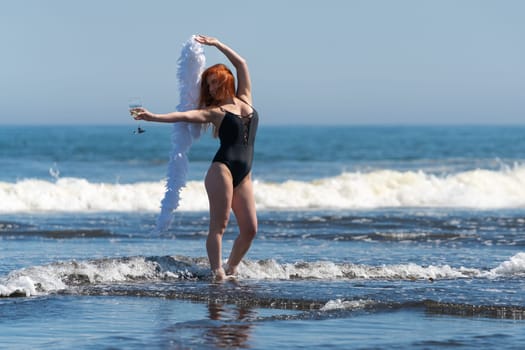 Smiling redhead woman in black one piece swimsuit stands ankle deep in waves of ocean and holding glass of wine in one outstretched hand and boa in other hand raised above head. Front view full length