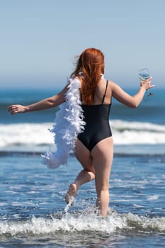 Playful and adventurous, an unrecognizable woman walks ankle deep into breaking waves of sea. With glass of wine in one hand and boa in other captures excitement and joy of living life to fullest