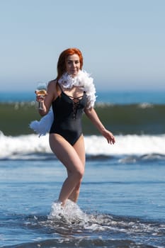 Gorgeous woman with fiery red hair is standing in refreshing ocean waves, holding glass of wine. She looks absolutely fabulous in her sleek black one-piece swimsuit and stylish boa wrapped around neck