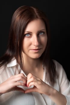 Portrait of woman showing heart shapes sign with both hands, looking at camera. Adult Caucasian ethnicity brunette female with gray eyes in white shirt on black background. Love at first sight concept