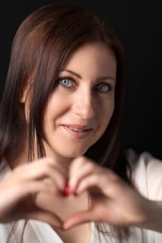 Brunette Caucasian ethnicity woman confesses in love, makes heart gesture with both hands and looking at camera, showing true feelings, happy expression, wears white shirt, poses on black background.