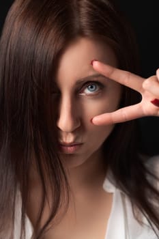 Portrait of serious adult woman holding two fingers near eye, showing victory or peace gesture and looking at camera. Sulking nice brunette with long hair covering one eye and outstretched lips.