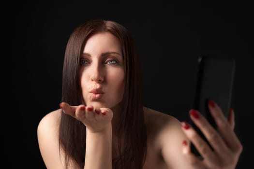 Woman blowing lips sending air kisses over palm and taking selfie to mobile phone camera. Charming 40s female with long hair flirting and looking at camera. Front view, studio shot on black background