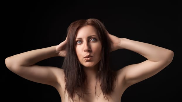 Studio portrait of beautiful and sexy woman with long hair and outstretched lips on black background. Sulking nice brunette model standing with raised hands behind her head and playfully looking away.