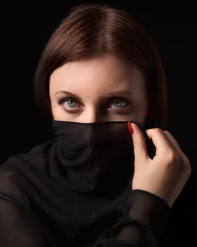 Shy brunette woman covered her nose and mouth or face with headscarf, looking at camera. Headshot of 40-year-old sexy woman with beautiful gray eyes and long hair. Portrait of lady on black background