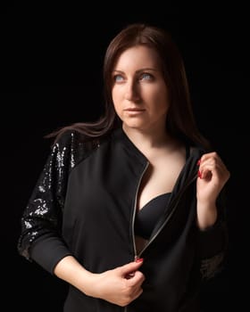 Shy brunette woman unzipping black jacket and showing bra, looking up away. Waist up portrait of beautiful and sexy 40-year-old lady with gray eyes and long hair on black background. Studio shot