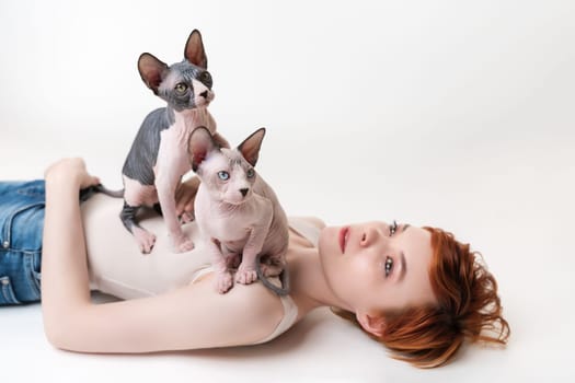 Two friendly Sphynx Hairless Cat, sitting on redhead young woman lying on back on white background. Beautiful woman with short hair wearing T-shirt and blue jeans. Part of series. Selective focus