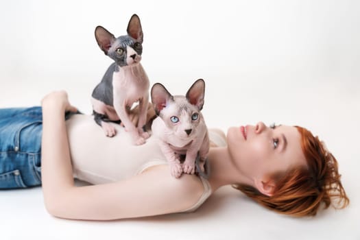 Two Canadian Sphynx Cat, sitting on young woman lying down on back on white background. Cute redhead woman with short hair wearing T-shirt and blue jeans. Part of series. Selective focus