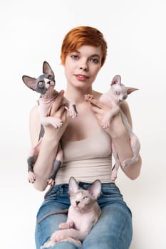 Redhead young woman holding two Sphynx Cat in hands and one kitten lying on her legs. Pretty woman with short hair in T-shirt and jeans looking camera, lying down on white background. Part of series