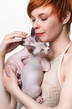 Beautiful redhead young woman gently hugging two purebred Canadian Sphynx Cat to her chest and looking down at domestic hairless kittens. Studio shot on white background. Part of series.