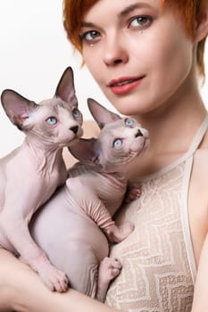 Cute redhead young woman gently hugging two domestic Sphynx Cat to her chest and looking at camera. Selective focus on friendly hairless kittens. Studio shot on white background. Part of series.