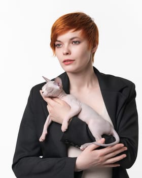 Young woman with short hair holding in hands sleeping Sphynx Hairless Cat blue mink and white color. Beautiful redhead woman dressed in black jacket on white background. Studio shot. Part of series.