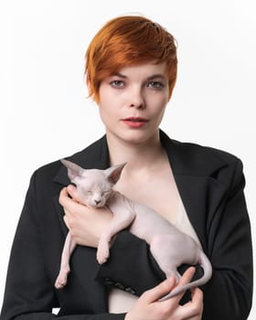 Bossy young woman dressed in black jacket holding in hands sleeping Sphynx kitten blue mink and white color. Pretty redhead woman with short hair on white background. Studio shot. Part of series.