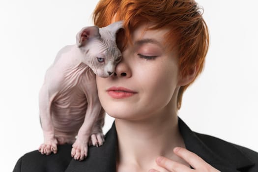 Redhead woman with closed eyes and playful kitten standing on shoulder, gently pressed against face. Selective focus on woman, shallow depth of field. Studio shot on white background. Part of series