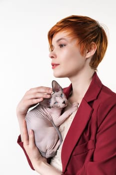 Hipster young redhead woman with short hair gently hugging to her chest Sphynx Hairless Cat. Beautiful woman looking away, dressed in red jacket. Studio shot on white background. Part of series.