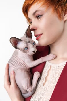 Hipster young redhead woman gently hugging Canadian Sphynx Cat. Selective focus on kitten, shallow depth of field. Cute woman dressed in red jacket. Studio shot on white background. Part of series.