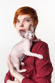 Canadian Sphynx Cat sitting on shoulder of hipster redhead young woman. Selective focus on foreground domestic hairless kitten, shallow depth of field. Studio shot, white background. Part of series