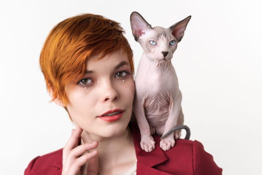 Portrait of beautiful redhead young woman with short hair, dressed in red jacket and playful cat sitting on her shoulder. Hipster woman looking at camera. Studio shot on white background. Part series.