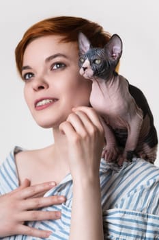 Sphynx kitten sitting on shoulder of happy redhead young woman. Selective focus on cat. Hipster female with short hair dressed striped white-blue shirt. Studio shot on white background. Part of series