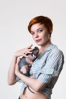 Redhead young woman gently hugging Canadian Sphynx Cat, looking at camera. Studio shot on white background. Portrait caucasian hipster with short hair dressed in striped white-blue shirt. Part series.