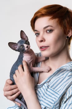 Beautiful redhead young woman holding Canadian Sphynx Cat in hands and looking away. Studio shot on white background. Hipster with short hair dressed in striped white-blue casual shirt. Part of series