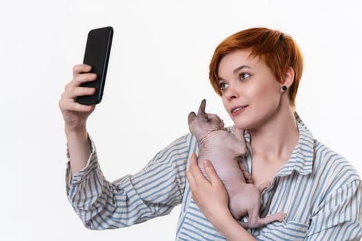 Redhead young woman hugging Sphynx Cat, taking selfie photo on cellphone, looking at telephone camera. Studio shot on white background. Hipster dressed in striped white-blue shirt. Part of series.