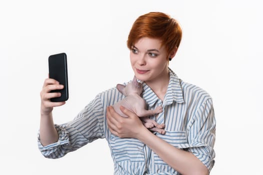Redhead young woman hugging sleeping domestic kitten, using her cell phone and taking selfie at smartphone camera. Studio shot on white background. Part of series. Facial expression, positive emotion