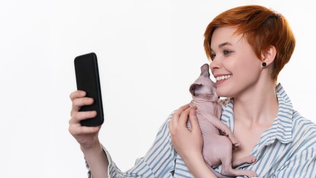 Smiling redhead young woman hugging Sphynx Cat, takes selfie photo on smartphone, looking at mobile phone camera. Beautiful hipster dressed in striped white-blue shirt on white background. Part series