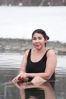 Busty curvy plus size young adult model in black one-piece bathing suit sitting in geothermal water in outdoors pool at balneotherapy health spa, hot springs resort having balneological properties