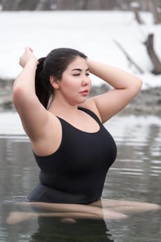 Large size young adult model with big breast in black one-piece bathing swimsuit sitting in outdoors pool at balneotherapy spa, hot springs resort. Woman raised hands straightens hair, looking away