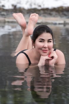 Plus size model in bathing suit lying down in geothermal mineral water in outdoor pool at balneotherapy health spa, hot springs resort having balneological properties. Woman looking at camera, smiling