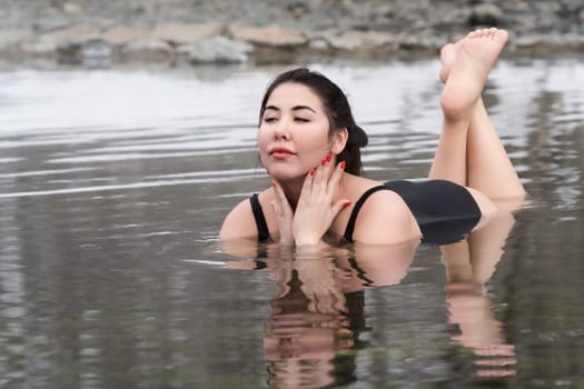 Full figured young adult model in black one piece bathing swimsuit lying down and relaxation in mineral water in outdoors pool at spa resort having balneological properties. Woman looking away
