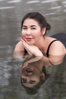 Extended sizes young woman in black swimming costume lying down geothermal mineral water in outdoors pool at balneotherapy spa, hot springs resort. Young adult model looking at camera. Body positive