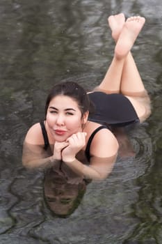 Overweight young woman in black one-piece bathing suit lying down, relaxation in geothermal mineral water in outdoors pool balneotherapy health spa, hot springs resort having balneological properties