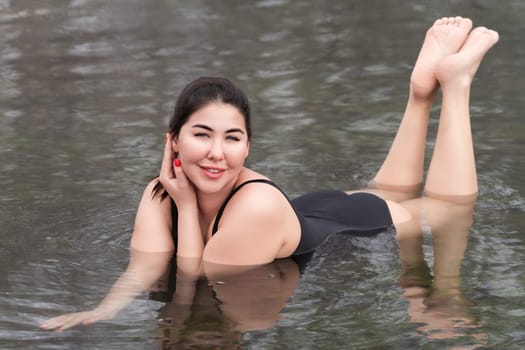 Happy outsize young woman in black one piece bathing swimsuit lying down and relaxation in mineral water in outdoors pool at spa, hot springs resorts. Woman looking at camera, smiling. Body positive