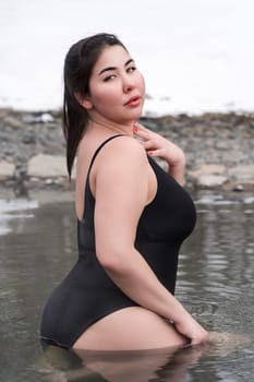 Busty curvy extended sizes young adult model in black one-piece bathing suit sitting in outdoors pool at balneotherapy spa. Side view of plus size figured woman with big breast looking over shoulder